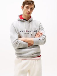 Jaunas vyras pilku Tommy Hilfiger gobtuvu su raudonu apykakle ir baltais džinsais, stovintis su sukryžiuotomis rankomis prieš baltą fono.