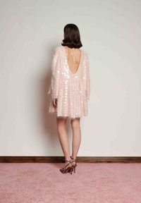 Kvinna i en ljusrosa, strukturerad klänning med djup rygg och leopardmönstrade högklackade remskor står på en rosa matta mot en enfärgad vägg.