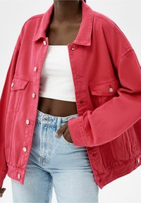 Veste en denim rose vif avec une coupe décontractée, grandes poches avant, boutons-pression en métal et coutures visibles. Portée sur un crop top blanc et un jean clair.