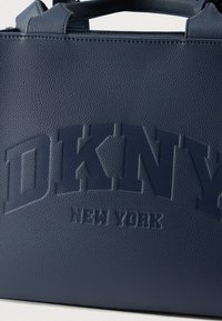 Tumesinise tekstuuriga nahast õlakott, mille peal on reljeefne "DKNY NEW YORK" logo ja kaks ülemist käepidet.