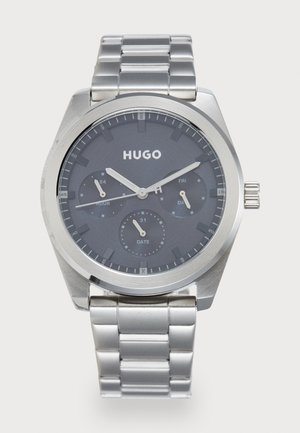 Silberne Metall-Armbanduhr mit dunkelgrauem Zifferblatt, drei Hilfszifferblättern für Stunde, Datum und Tag sowie „HUGO“-Logo unter 12 Uhr.