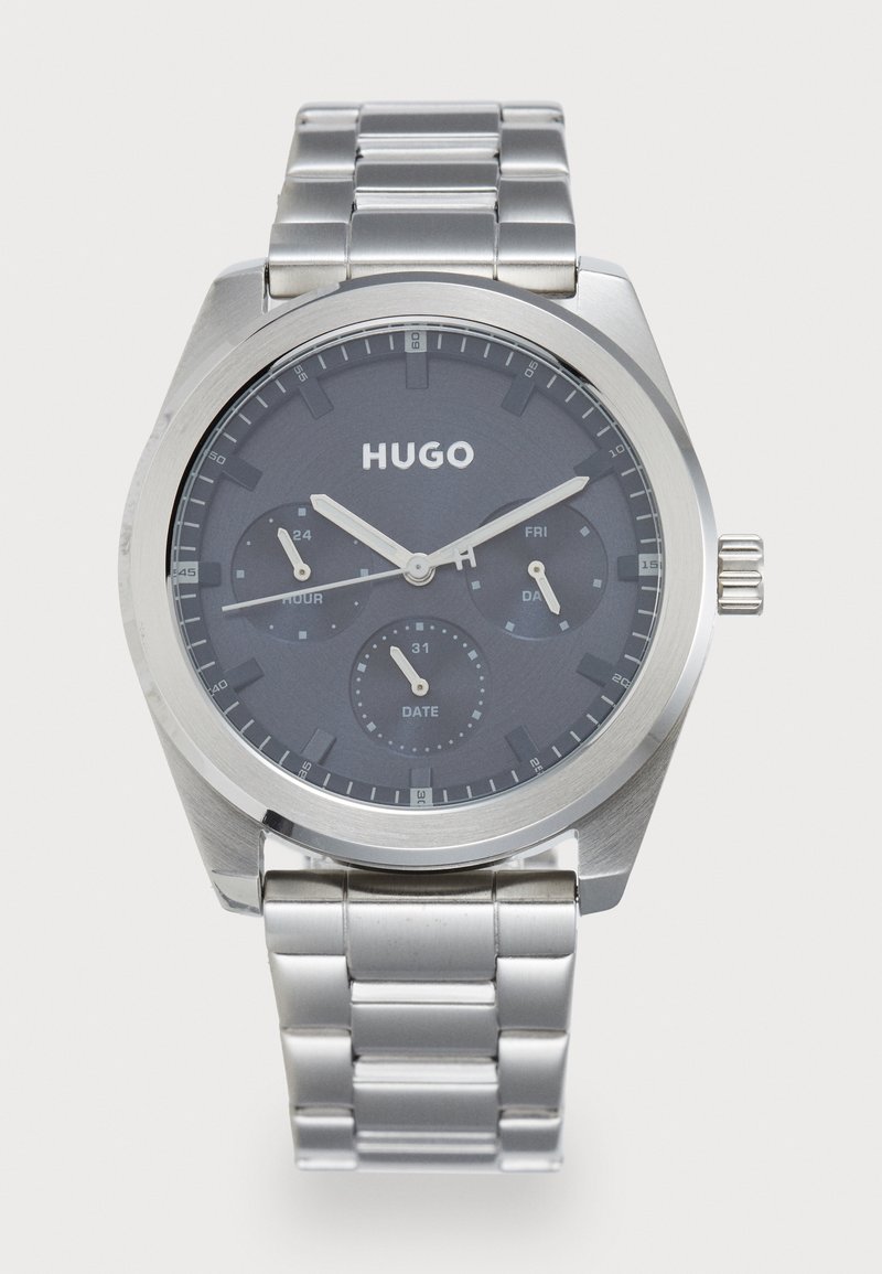 Montre-bracelet en métal argenté avec cadran gris foncé, trois sous-cadrans pour l'heure, la date et le jour, et logo "HUGO" sous les 12 heures.