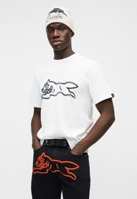 Άσπρο βαμβακερό μπλουζάκι με μαύρο γραφικό ενός τρέχοντος σκύλου, συνδυασμένο με σκούρο τζιν παντελόνι που παρουσιάζει έναν πορτοκαλί τρέχοντα σκύλο.