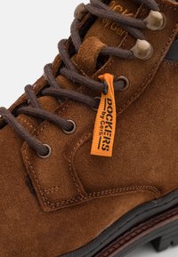 Stivale alla caviglia in suede marrone con design con lacci, superficie testurizzata, occhielli in metallo e etichetta arancione del marchio. Accento nero sul lato. Suola spessa.