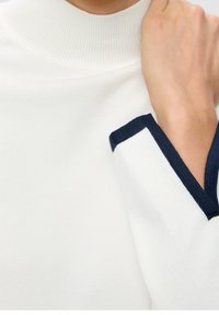 Närbild av en hand som rör vid nacken, klädd i en vit tröja med hög krage och marinblå kant på ärmsluten.