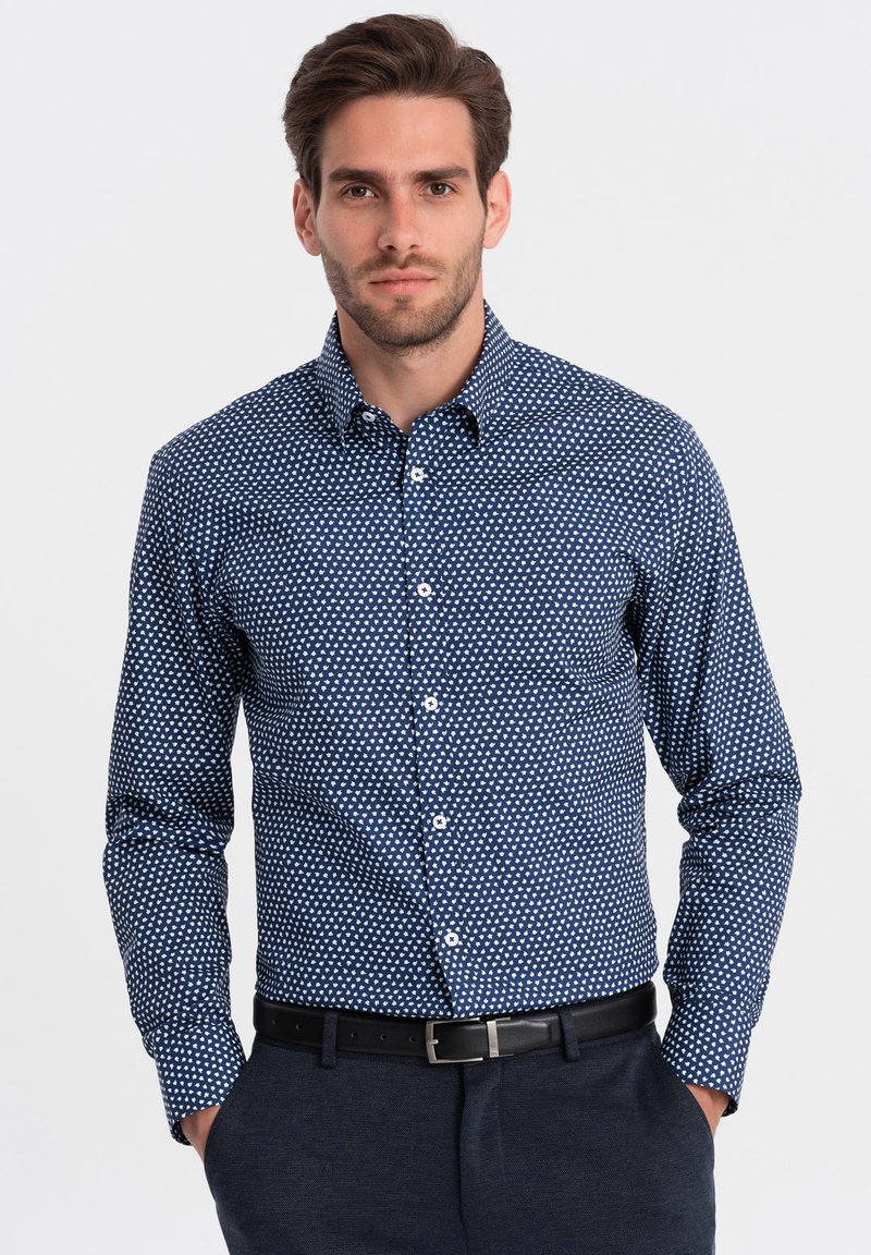 Camicia blu navy a maniche lunghe con bottoni, decorata con pois bianchi, dotata di un taschino sul petto e colletto a punta; abbinata a pantaloni scuri con cintura.
