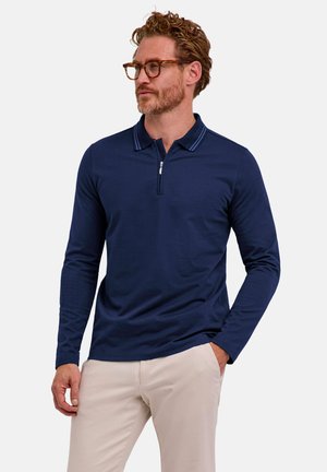 Mann mit lockigem Haar und Brille, der ein dunkelblaues langärmliges Poloshirt mit Reißverschluss und beigen Hosen trägt, steht mit einer Hand in der Tasche.