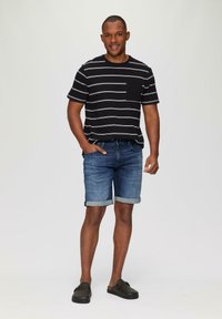 Zwart gestreept T-shirt, blauwe denimshorts met opgerolde zomen, en donkere instapschoenen. Effen achtergrond, casual outfit.