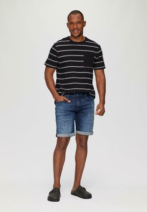 Zwart gestreept T-shirt, blauwe denimshorts met opgerolde zomen, en donkere instapschoenen. Effen achtergrond, casual outfit.