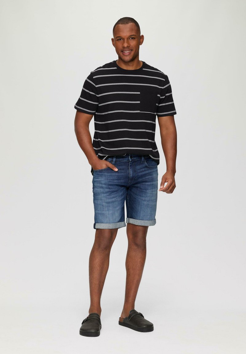 Zwart gestreept T-shirt, blauwe denimshorts met opgerolde zomen, en donkere instapschoenen. Effen achtergrond, casual outfit.