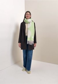 Groen sjaal met patroon en franjes, gedragen over een crème trui en een bruine jas, gecombineerd met blauwe wijde jeans en lichte sneakers.