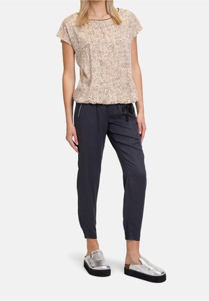 Frau trägt eine beige gepunktete Kurzarmbluse, marineblaue Jogginghose mit Kordelzug und silberne Slip-on Plateau-Schuhe, steht auf weißem Hintergrund.