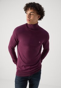 Pull encolure montant bordeaux en tissu doux, avec un design ajusté et des poignets côtelés. Petit logo sur la poitrine, associé à un jean foncé.