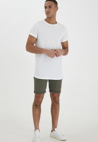 Hvitt bomull T-skjorte med rullede ermer, kombinert med olivengrønne shorts og hvite sneakers. Modellen har en tatovering på beinet.