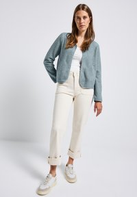 Giacca in pile azzurro chiaro con finitura textured, design cropped, scollo aperto, abbinata a pantaloni bianchi a vita alta e sneaker color crema.