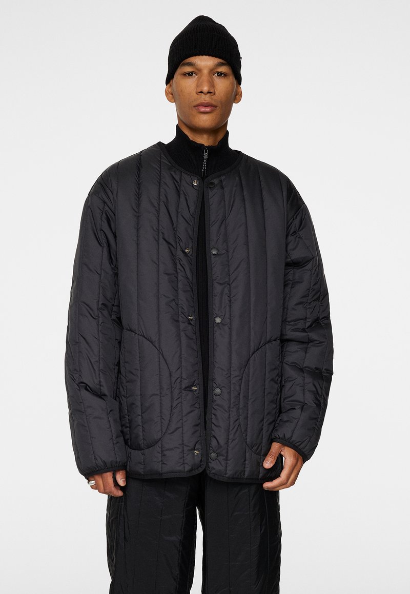 Svart quiltad jacka med rund halsringning, tryckknappsstängning och subtila sömnadsdetaljer. Bärs över en svart zip-up topp.