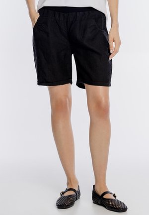 Kobieta ubrana w czarne szorty z elastycznym pasem i podwiniętymi nogawkami oraz czarne plecione baleriny typu Mary Jane, stojąca z rękami w kieszeniach.