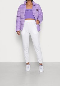 Lila Pufferjacke mit hohem Kragen, kombiniert mit einem lilafarbenen Croptop und weißen Jogginghosen. Weiße Sneaker vervollständigen das Outfit.
