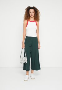 Wit mouwloos shirt met rode bandjes, groene broek met wijde pijpen en witte sneakers. Een zilveren handtas vasthoudend, staand tegen een effen achtergrond.