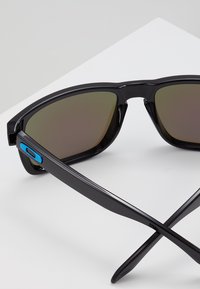 Sunglasses with a black plastic frame, brown gradient lenses, and a blue accent on the arm. The design features sleek, angular lines.