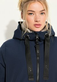 Marinfärgad vattentät jacka med silvrigt foder på huvan, svart dragkedja och justerbara svarta remmar. Har en minimalistisk design och en jämn textur.