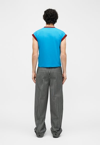 Homme aux cheveux bouclés portant une chemise à manches courtes bleue avec un bord rouge et un pantalon large à rayures verticales noir et blanc, debout, tourné de dos.