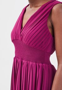 Robe sans manches magenta avec un corsage plissé, une ceinture large smockée et une jupe plissée fluide sur une personne à la peau foncée.