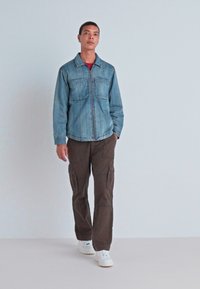 Joven caminando con pantalones cargo marrones, chaqueta de denim azul abrochada, camiseta roja debajo y zapatillas blancas sobre un fondo liso.