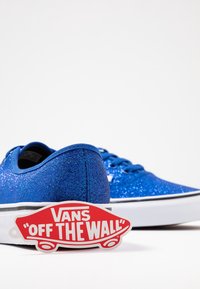 Sneakers blu con una finitura luccicante, suola bianca e striscia nera, dotati di un'etichetta rossa e bianca "Vans Off The Wall".