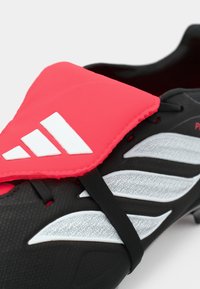 Μαύρο ποδοσφαιρικό παπούτσι με ένα φωτεινό ροζ πάνελ, λευκό λογότυπο Adidas και υφή με λεπτομέρειες. Διαθέτει ανακλαστικές λεπτομέρειες και ιμάντα ασφαλούς κλεισίματος.