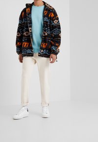 Chaqueta de forro polar en negro, naranja, azul y un patrón tribal blanco; sudadera azul claro; pantalones crema; zapatillas blancas con detalles negros.