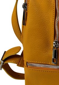 Geel leren rugzak met een gestept textuur, zilveren ritsen en een verstelbare schouderband. Heeft een zijvak met een klepafsluiting.