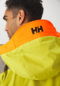 Chaqueta con capucha de alta visibilidad en tela amarilla y naranja. Presenta el logotipo negro "HH" en el cuello, textura suave y diseño estructurado.