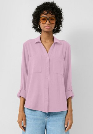 Kobieta o kręconych włosach, nosząca duże bursztynowe okulary przeciwsłoneczne, kolczyki koła, jasnofioletową koszulę na guziki i jasnoniebieskie dżinsy na jednolitym tle.