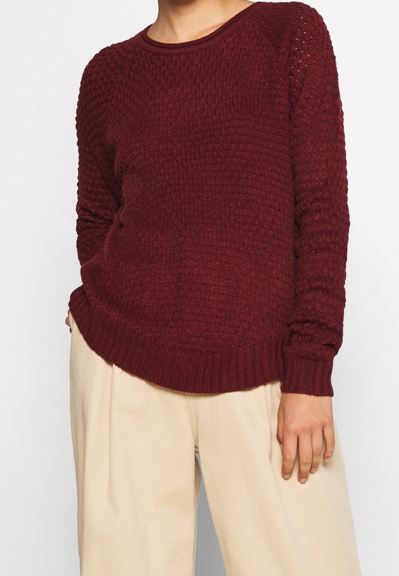 Personne portant un pull en maille rouge foncé texturé et un pantalon beige, debout devant un fond clair neutre.