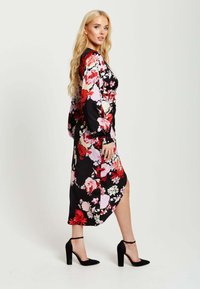 Floral dress features a black background with red and pink flowers, long sleeves, side slit, and a fitted silhouette. Black high-heeled shoes.