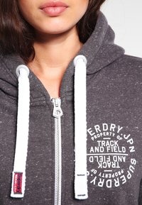 Sudadera con capucha gris con cremallera y cordones blancos. Presenta un estampado de logo y cremallera plateada. Tejido suave con una ligera textura y un diseño deportivo.