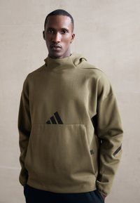 Felpa con cappuccio di colore verde oliva, dotata di tasca frontale. Presenta il logo Adidas nero e dettagli neri sulle maniche. Tessuto morbido, vestibilità rilassata.