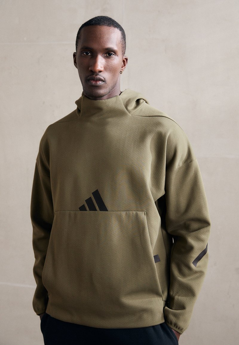 Felpa con cappuccio di colore verde oliva, dotata di tasca frontale. Presenta il logo Adidas nero e dettagli neri sulle maniche. Tessuto morbido, vestibilità rilassata.