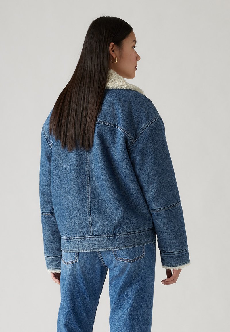 Veste en denim avec un col en sherpa, couleur bleue, tissu texturé, coupeLoose et détails cousus ; associée à un jean bleu assorti.