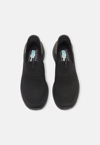 Zapatillas negras tipo slip-on con una parte superior de malla texturizada y bandas elásticas. Tienen una punta redondeada y una suela acolchada. Logo "SKECHERS" en la plantilla.