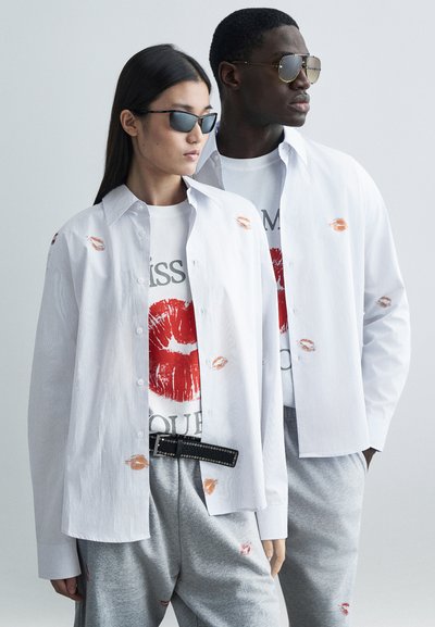 Biała koszula na guziki z nadrukiem pocałunków, noszona na t-shircie z grafiką czerwonych ust. Szare spodnie dresowe i czarny pasek z ćwiekami dopełniają stylizację.