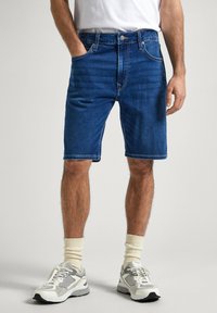 Pantalones cortos de mezclilla en azul oscuro con un corte clásico, cinco bolsillos, detalles de costura claros y cierre de cremallera, combinados con zapatillas gris y blancas.