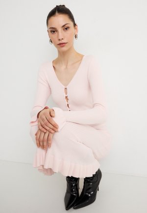 Jasnoróżowa prążkowana sukienka dzianinowa z dekoltem w serek, guzikami z przodu, rozkloszowanymi długimi rękawami oraz falbaną na dole, zestawiona z czarnymi, spiczastymi botkami za kostkę.