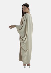 Beige oversized kaftan met gedrapeerde mouwen en een gladde textuur, met een ronde halslijn, reikt tot halverwege de kuit en is gecombineerd met witte blokhakken.