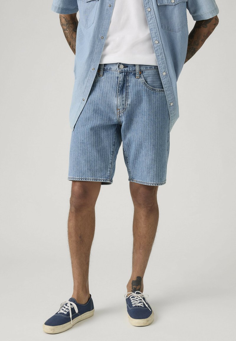 Hombre con pantalones cortos de mezclilla azul claro, camiseta blanca, camisa azul claro desabrochada y zapatillas azul marino, de pie con las manos detrás de la espalda.