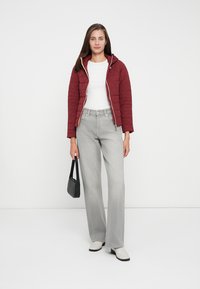 Veste bordeaux matelassée avec une capuche, associée à un haut blanc et un jean large gris clair. Sac à main noir, bottines blanches.