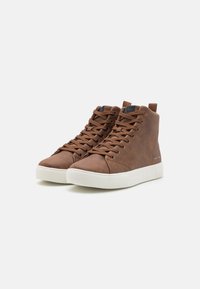 Brune høyhælte sneakers laget av glatt lær med teksturert finish, med en hvit gummisåle og matchende brune snørebånd.