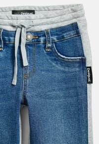 Džínové jogger kalhoty s modrým ombresením, šedým pasem, šňůrkami a logem. Obsahují tradiční kapsy a jemné prošívání.