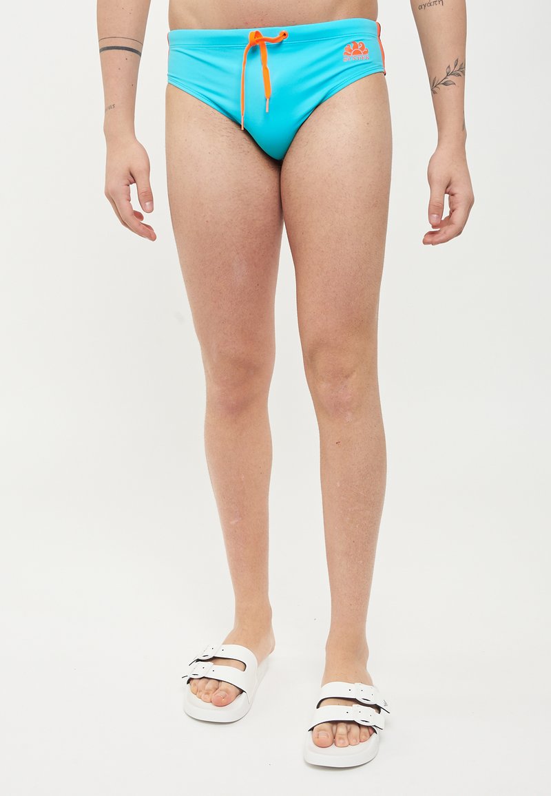 Bañador azul aqua con un cordón naranja, con un detalle de logo. Combinado con sandalias blancas de deslizar con acentos negros.